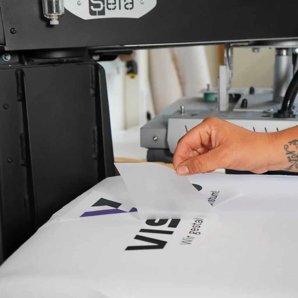Eine Hand zieht die Trägerfolie von einem frisch gepressten Shirt mit VISARO Logo ab