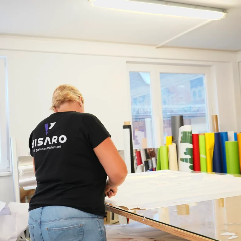 Mitarbeiterin im VISARO Shirt beim Zuschnitt am Arbeitstisch, dahinter Folienrollen
