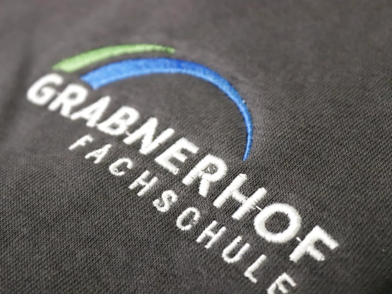 Gestickter Schriftzug „Grabnerhof Fachschule“ auf grauem Fleece