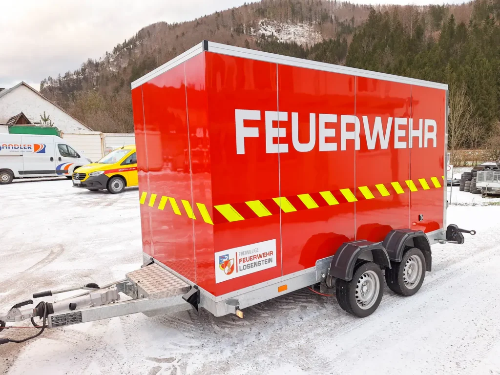 Roter Feuerwehranhänger mit weißer Aufschrift und gelb-roter Warnmarkierung im Schnee