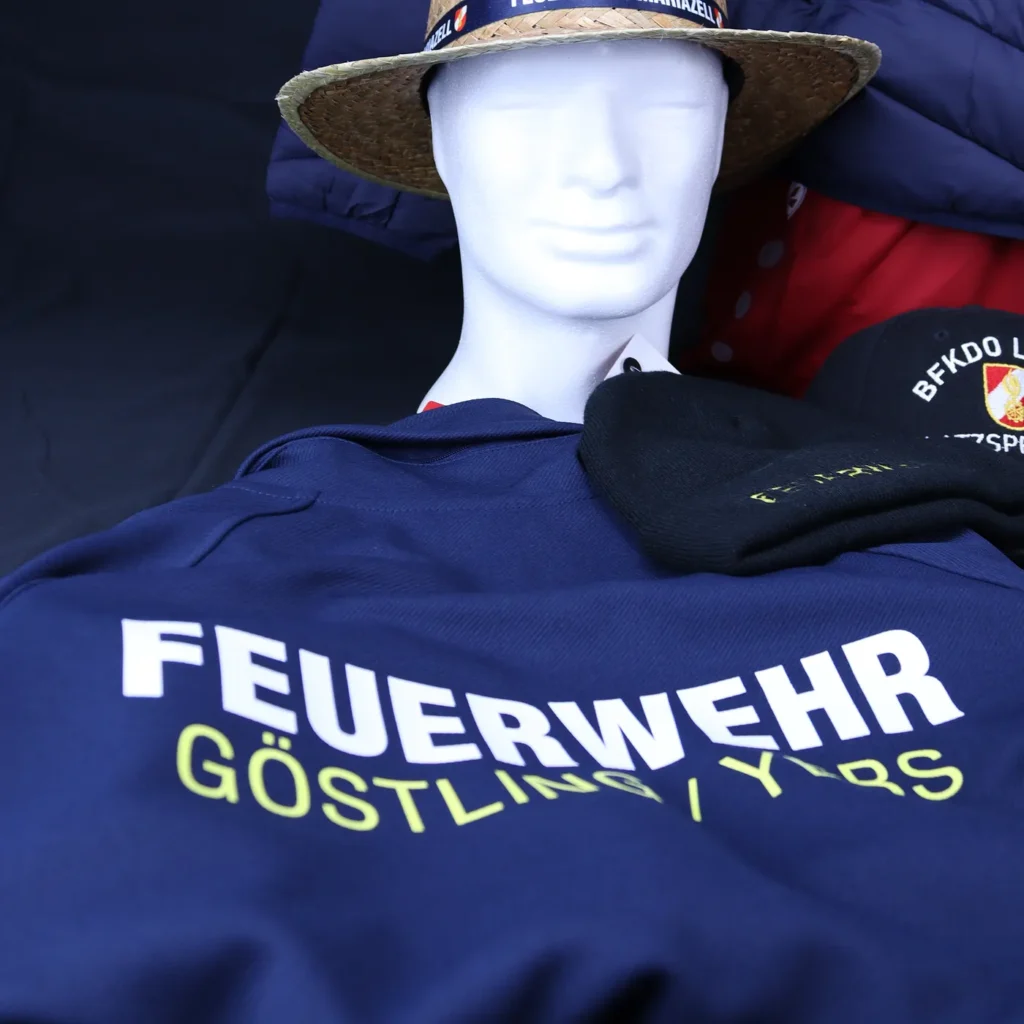 Dunkelblauer Hoodie mit Rückendruck „Feuerwehr Göstling/Ybbs“, dahinter Strohhut und bestickte Haube
