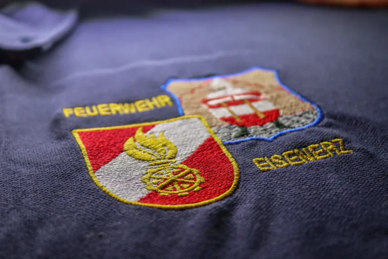 Gestickte Wappen „Feuerwehr“ und „Eisenerz“ auf dunklem Stoff